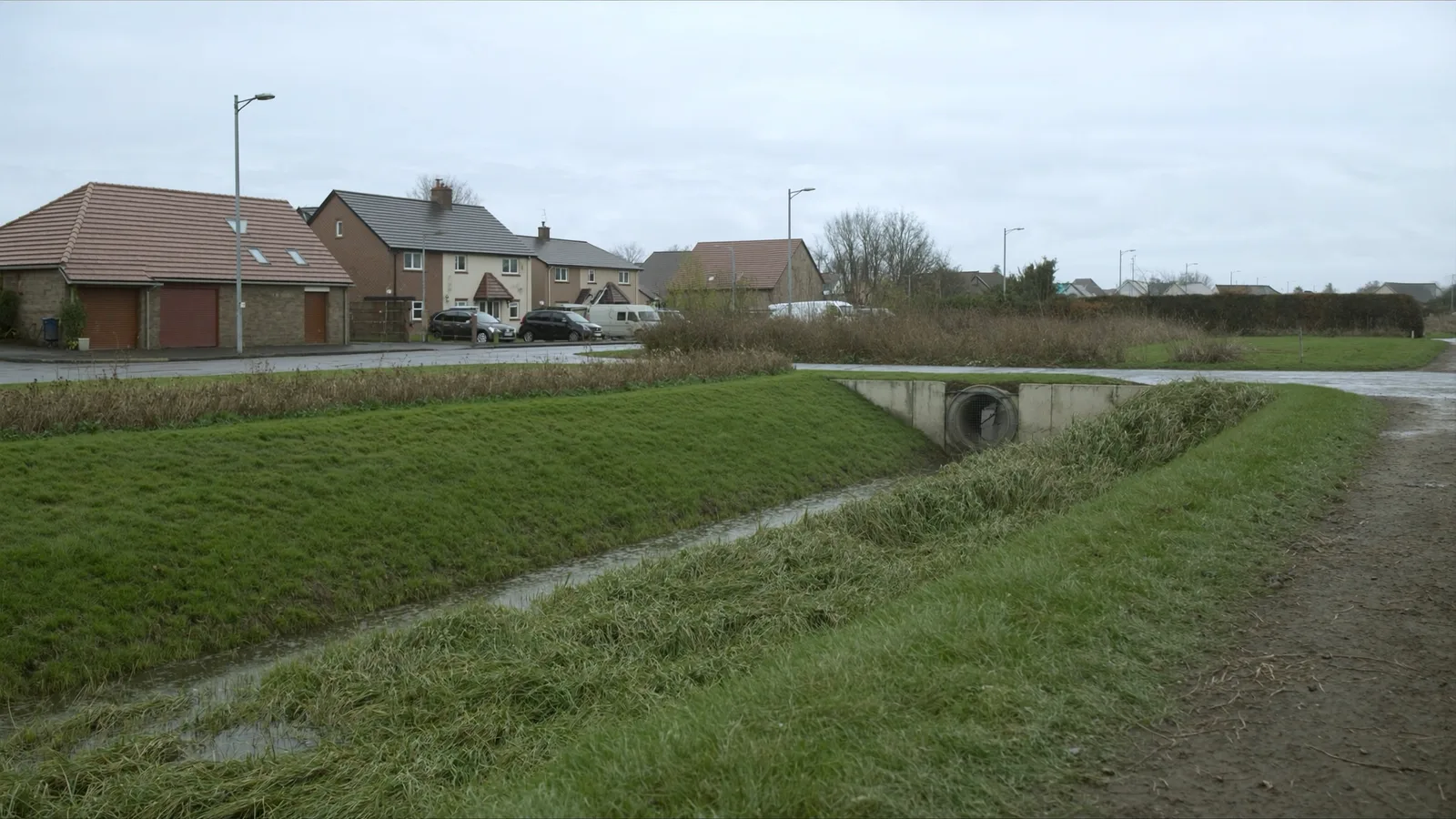 A grassed drainage swale and concrete headwall outfall alongside a newly built residential access road after rain.
